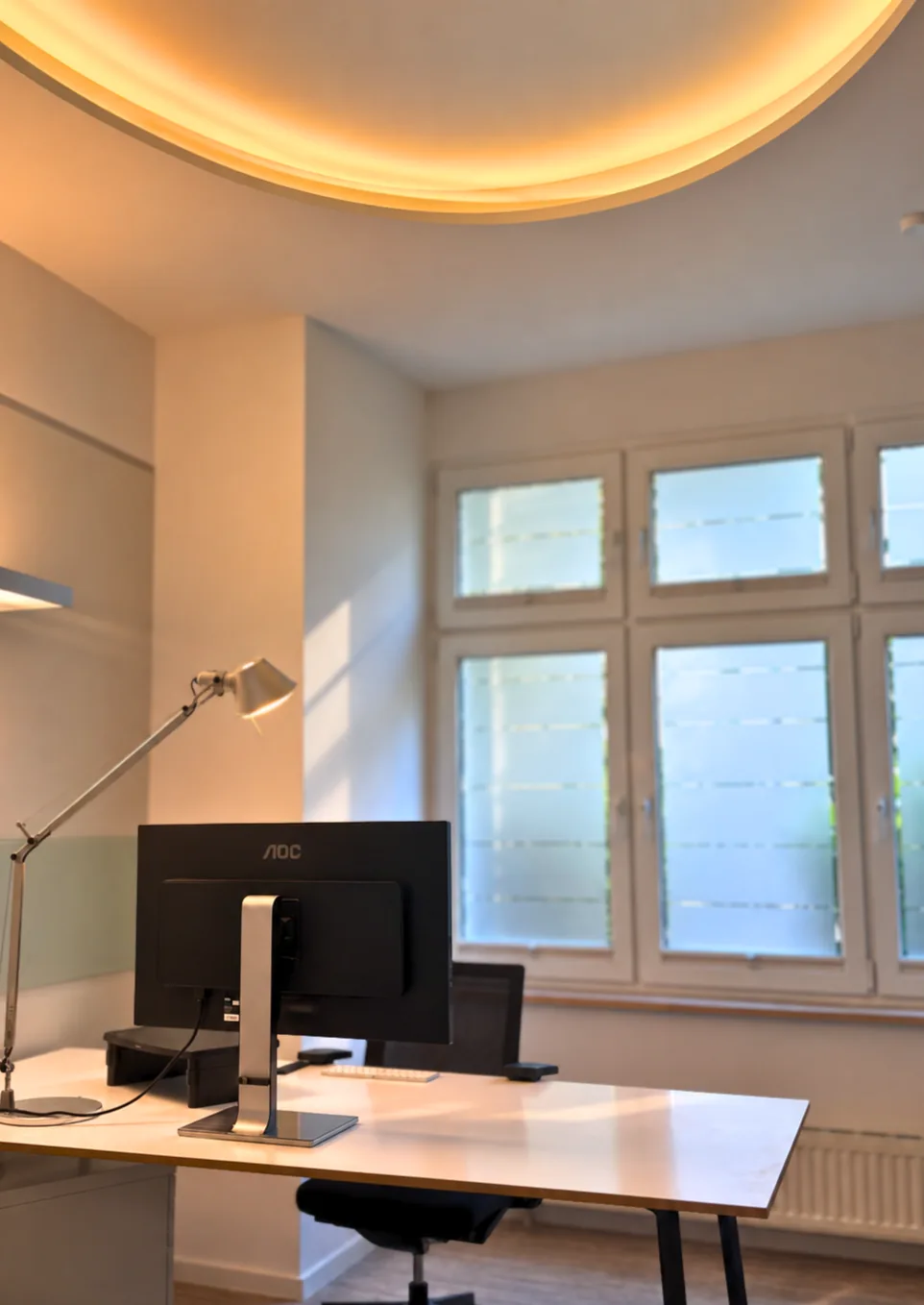 Das private Büro bei Flexovo Tempelhof: höhenverstellbarer Schreibtisch mit Monitor und Architektenlampe, Fenster mit Tageslicht und indirekte Deckenbeleuchtung
